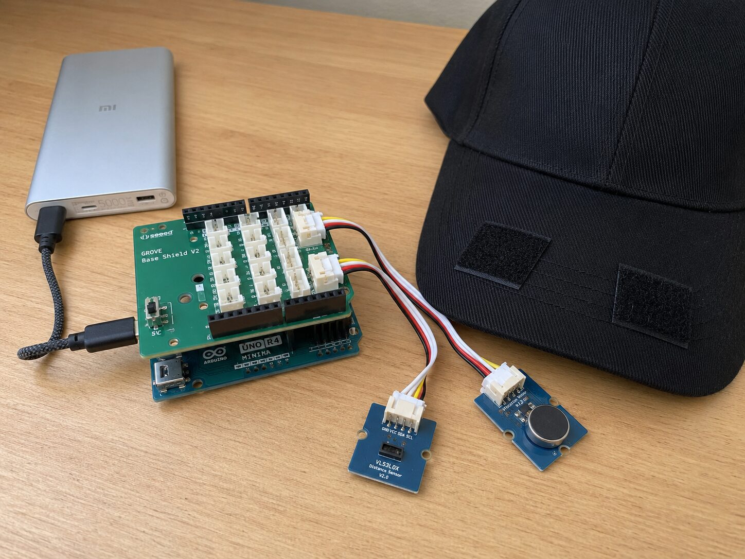 Concept mockup of the bench build: Arduino UNO with a Grove base shield, plug-in sensor and vibration motor modules, a power bank, and a black cap with Velcro strips on a wooden desk.