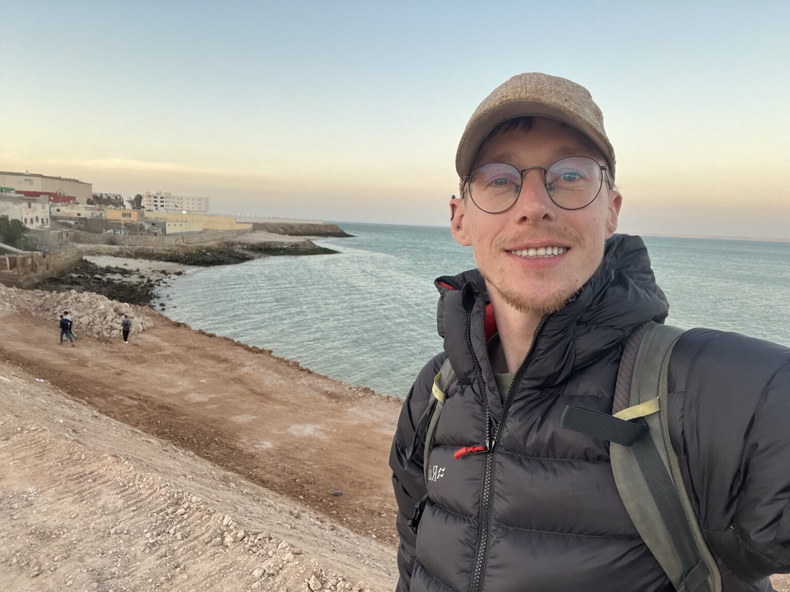 Josiah smiling in a cap and down jacket on a coastal path, sea and low buildings behind him.