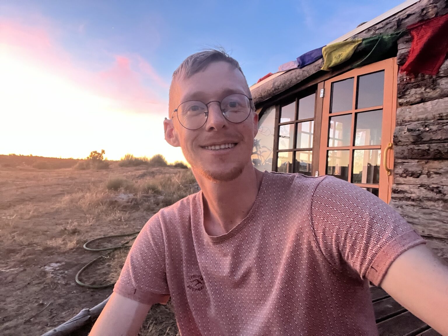 Josiah at sunset outside a rustic wooden cabin in rural Portugal, dry fields and an orange sky behind him.